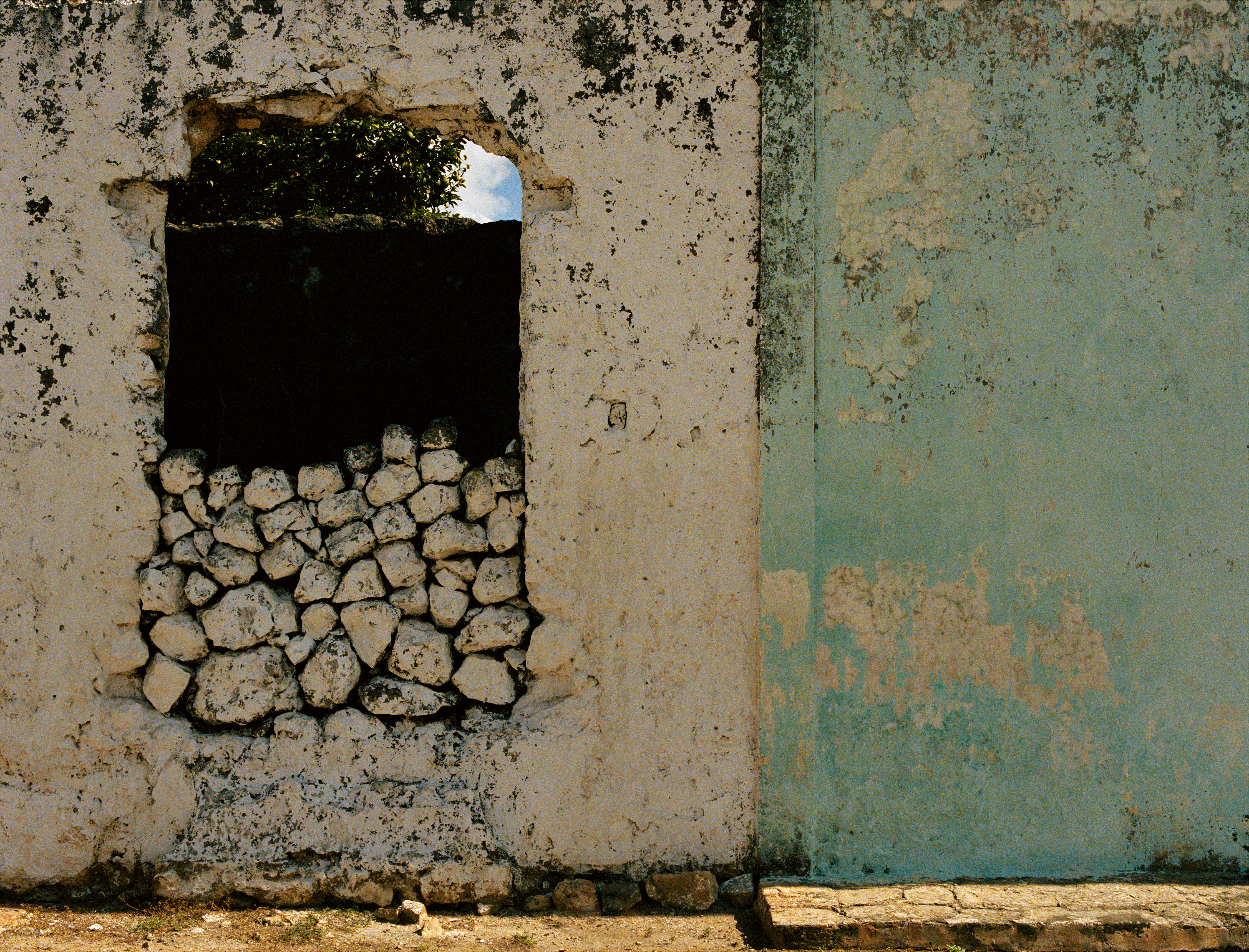 The photograph shows a house wall that is divided into two sections: on the left, it is beige, while on the right it is a vibrant turquoise. At the upper edge of the wall, the plaster is crumbling away, creating a rough, irregular texture. There is a window in the middle of the beige wall, in which stones are stacked at the bottom. 