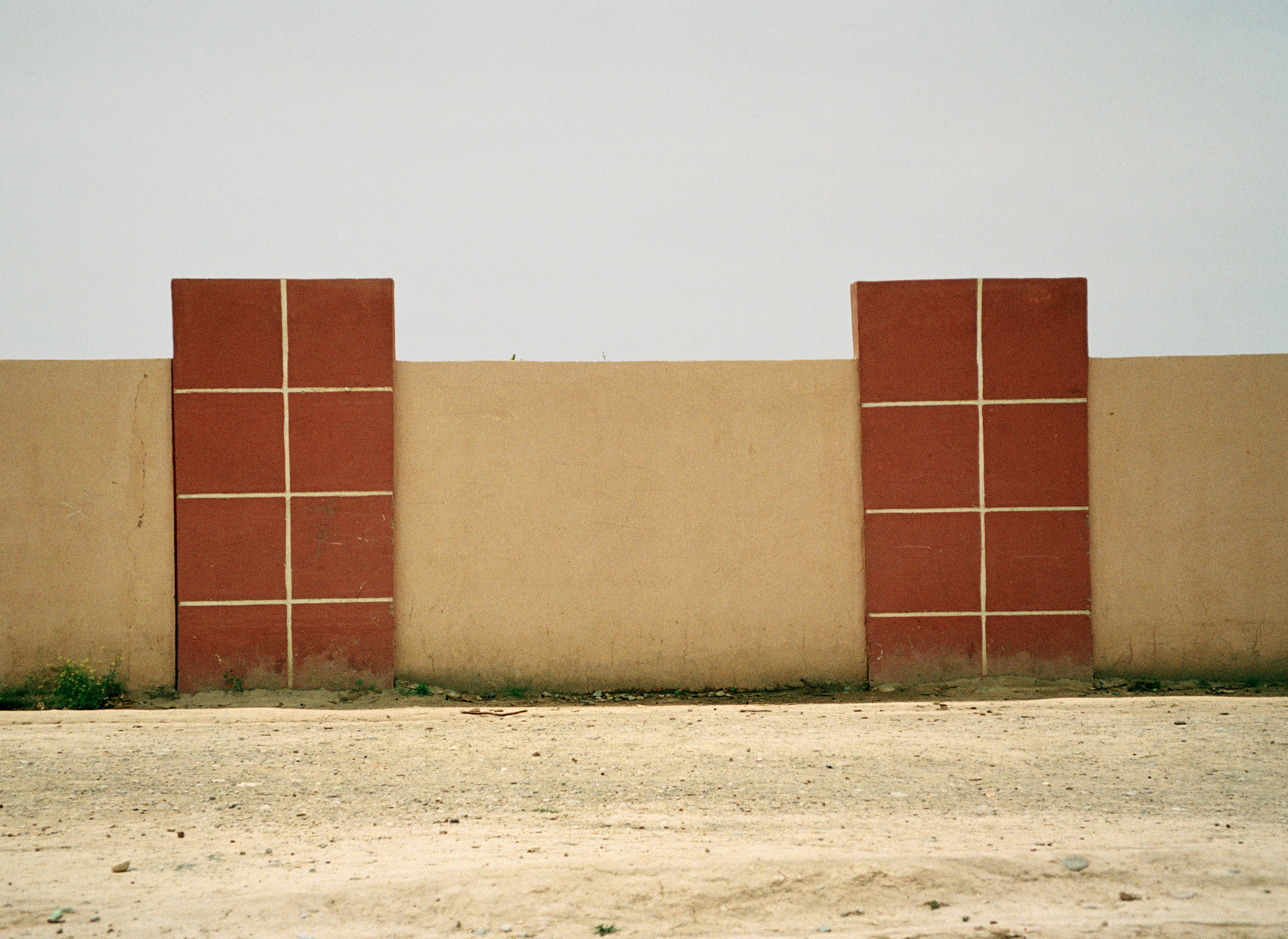 The photograph shows a strictly geometric composition reminiscent of Sean Scully's paintings. In the lower third of the picture is a beige floor, presumably sand. Above it rises an ochre-yellow and white-painted wall, on the left and right of which there is a rust-brown rectangle divided into eight smaller squares by fine yellow-beige lines. Despite the photographic image, the picture almost looks like an abstract painting due to its clear surfaces, lines and color contrasts - only at the lower edge do green plants slightly break the strict order.