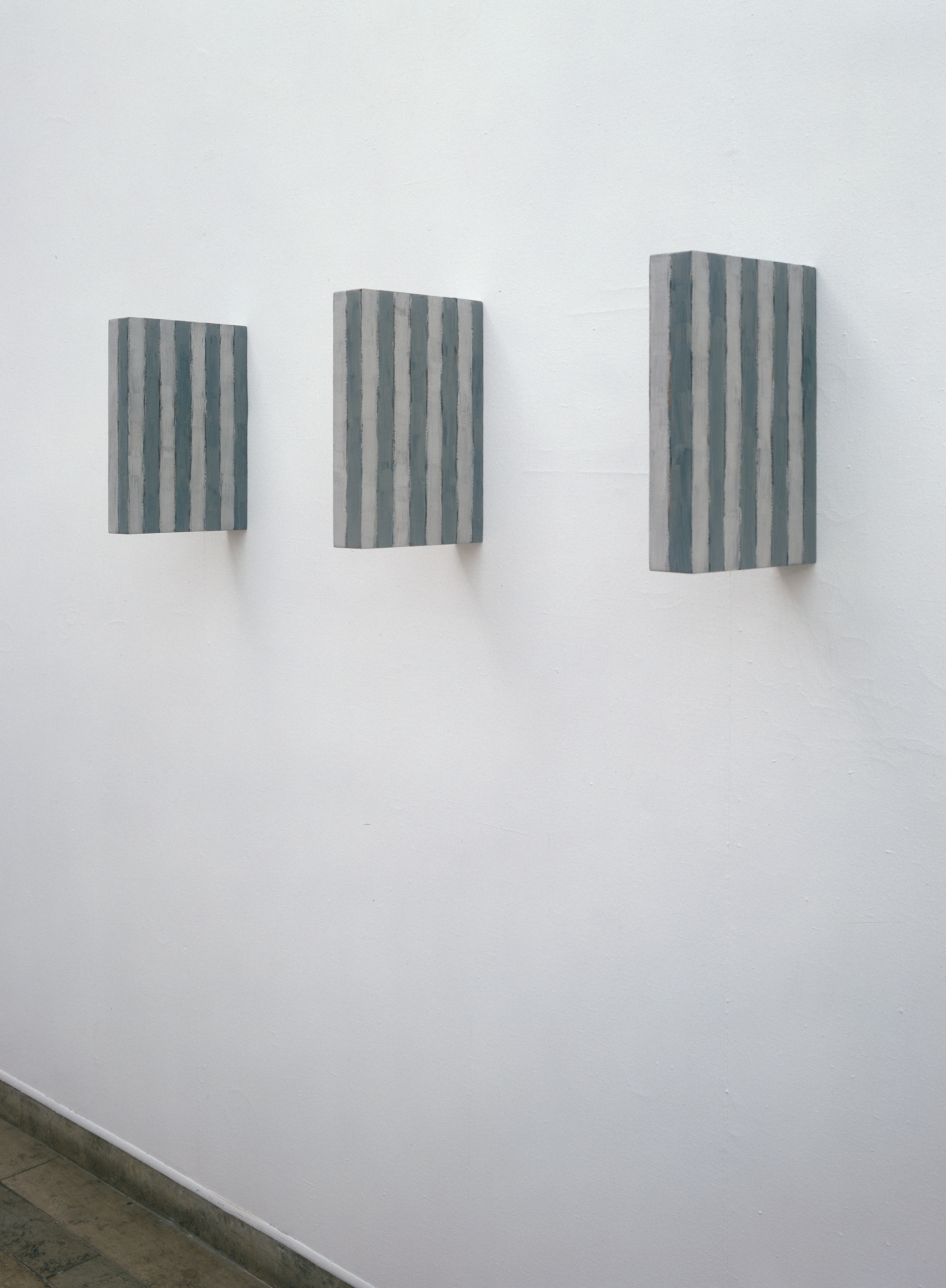 The work consists of three portrait-format aluminum panels that hang evenly next to each other on the wall. Each panel is covered with vertical stripes in blue-grey and white that run evenly and parallel. The colors alternate at clear, even intervals and the stripes are arranged at an equal width. The panels are positioned at an even distance from each other, creating a structured arrangement in the room.