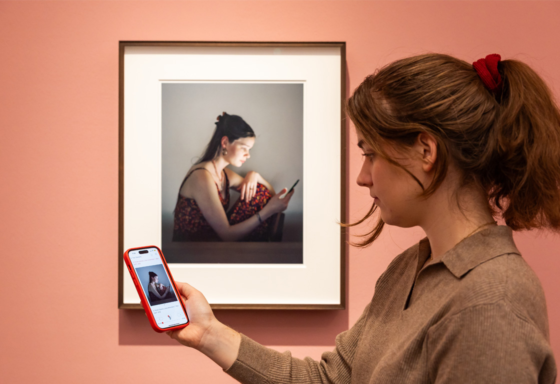 Eine Frau hält ein Handy auf dem der BKF-Audioguide läuft vor der Fotografie einer jungen Frau, die in einem dunklen Raum sitzt und auf ihr Smartphone blickt.