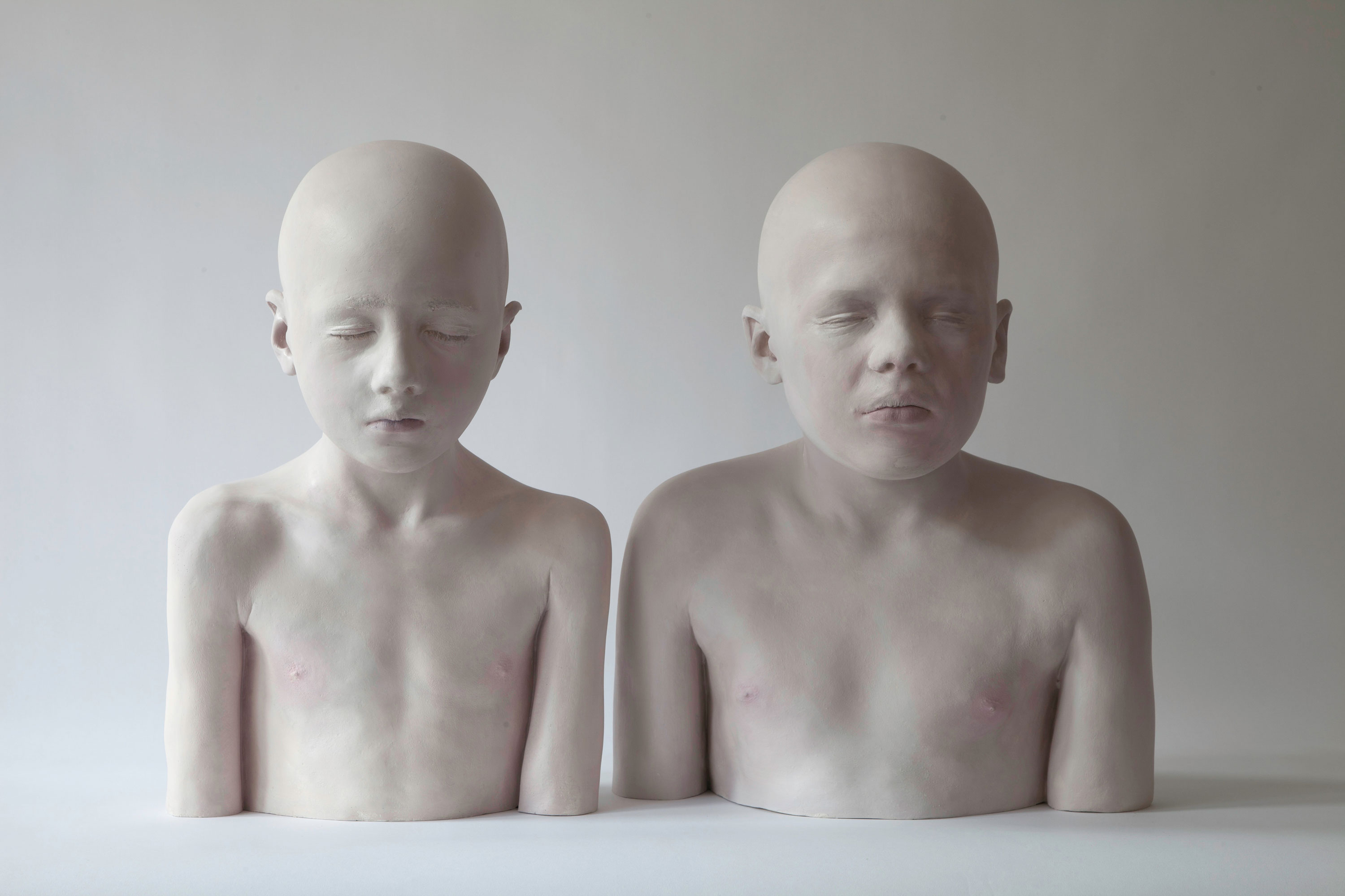 Das Bild zeigt zwei realistische Skulpturen von Kindern, die bis zur Brust dargestellt sind. Beide Skulpturen haben geschlossene Augen, eine blasshäutige Oberfläche und eine Glatze. Die Figuren stehen nebeneinander vor einem neutralen, hellen Hintergrund. 