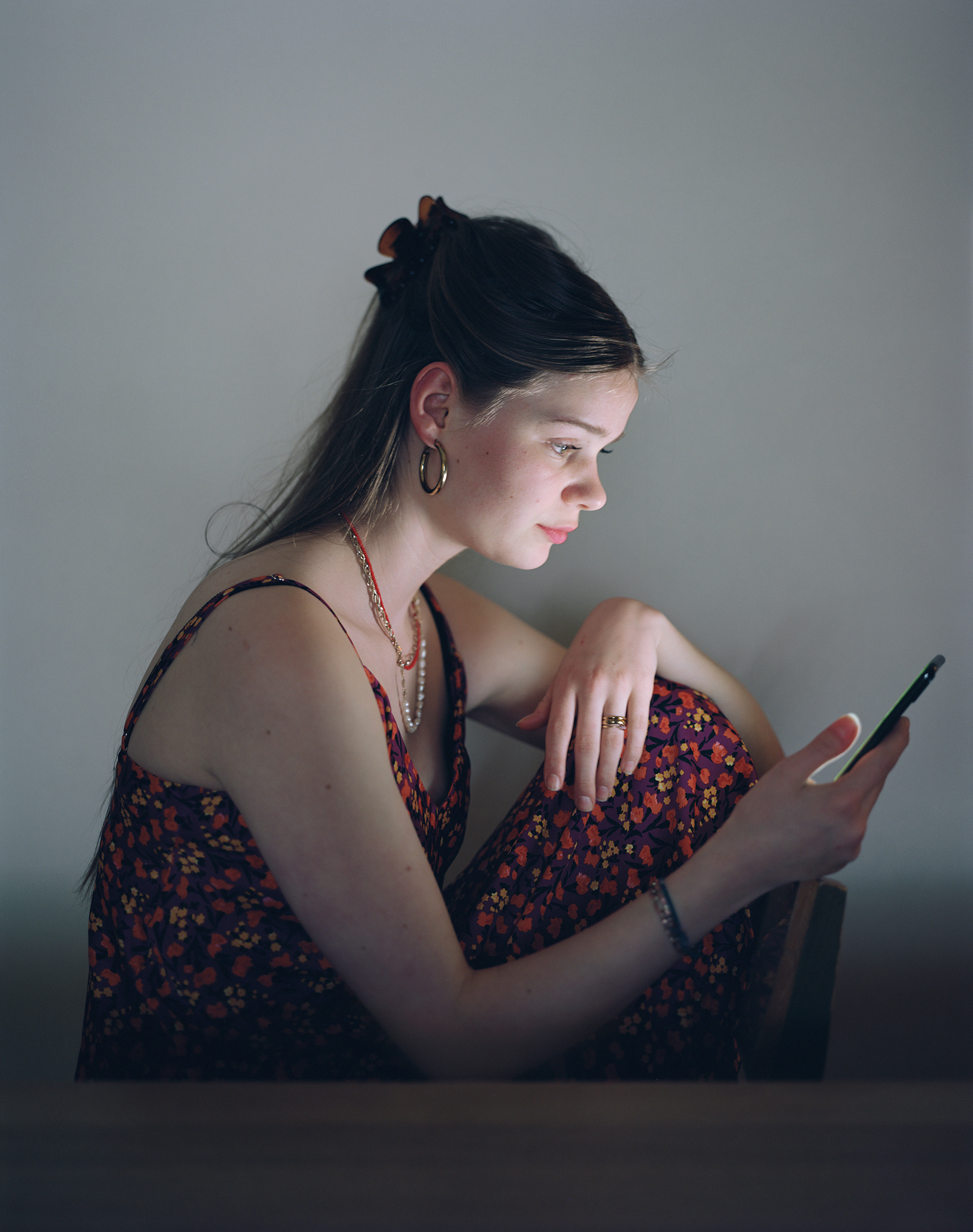 Das Foto zeigt eine junge Frau, die in einem dunklen Raum sitzt und auf ihr Smartphone blickt. Sie trägt ein ärmelloses Kleid mit einem Blumenmuster und hat ihre langen Haare mit einer Spange zurückgesteckt. Ihr Gesicht wird sanft vom Bildschirmlicht beleuchtet, was einen konzentrierten Ausdruck hervorhebt. Der Hintergrund ist schlicht und dunkel, wodurch der Fokus auf die Frau und ihre Beschäftigung mit dem Smartphone gerichtet ist.