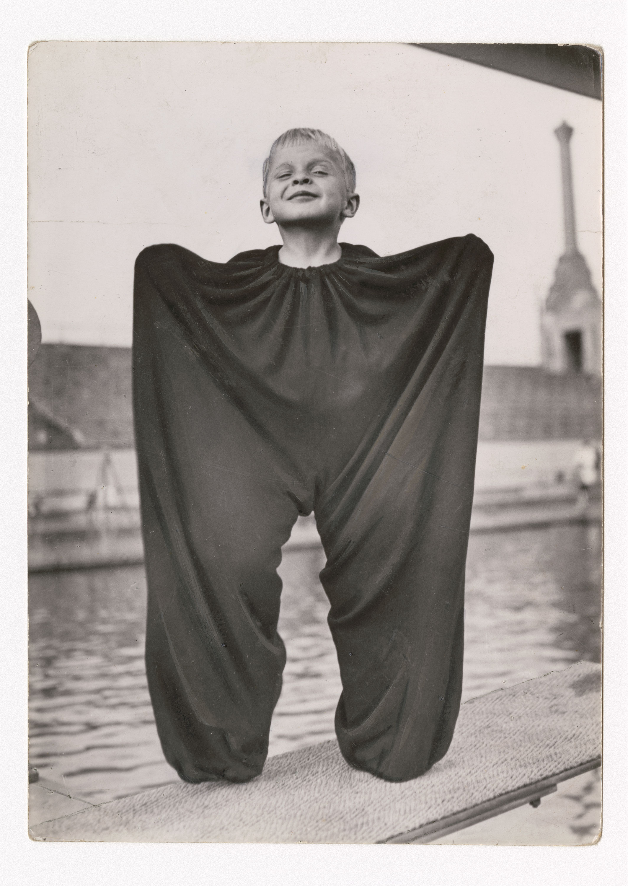 Das Foto zeigt einen Jungen, der in einer dunklen, übergroßen Hose steht, die aus dem Nachlass seines Vaters stammt. Die Hose ist so groß, dass sie ihm bis zum Hals reicht. Sie wirkt fast wie Flügel, da der Junge demonstrativ seine Arme ausbreitet, um sie an der Taille hochzuhalten. Er steht vor einer unscharfen Kulisse mit Wasser und einem Gebäude im Hintergrund. Der Ausdruck des Jungen ist fröhlich und unbeschwert.