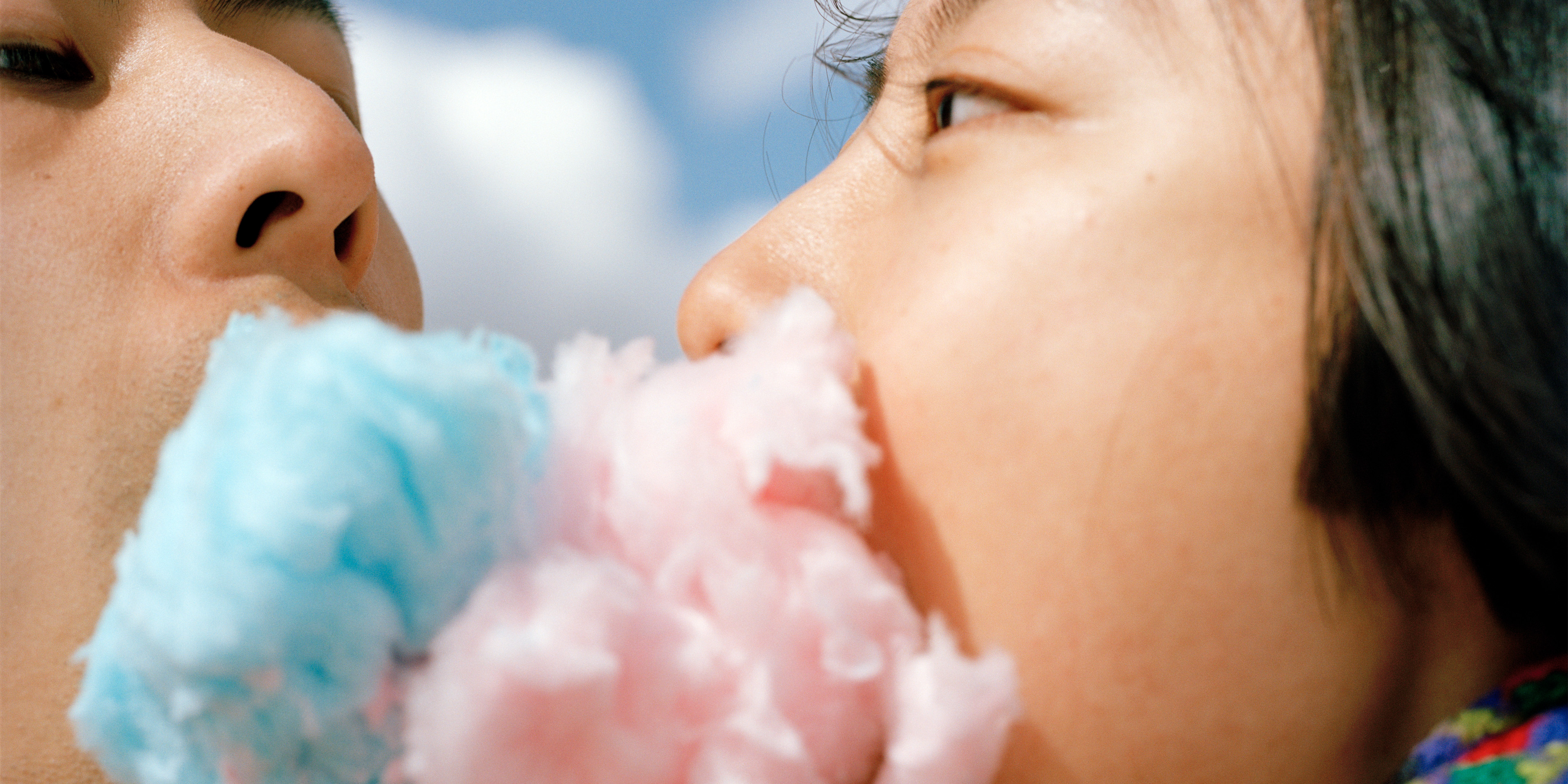 Zwei Personen, die sich nah gegenüberstehen. Die rechte Person hat blaue Zuckerwatte, die linke Person Zuckerwatte im Mund. Die Zuckerwattebäuschchen berühren sich.