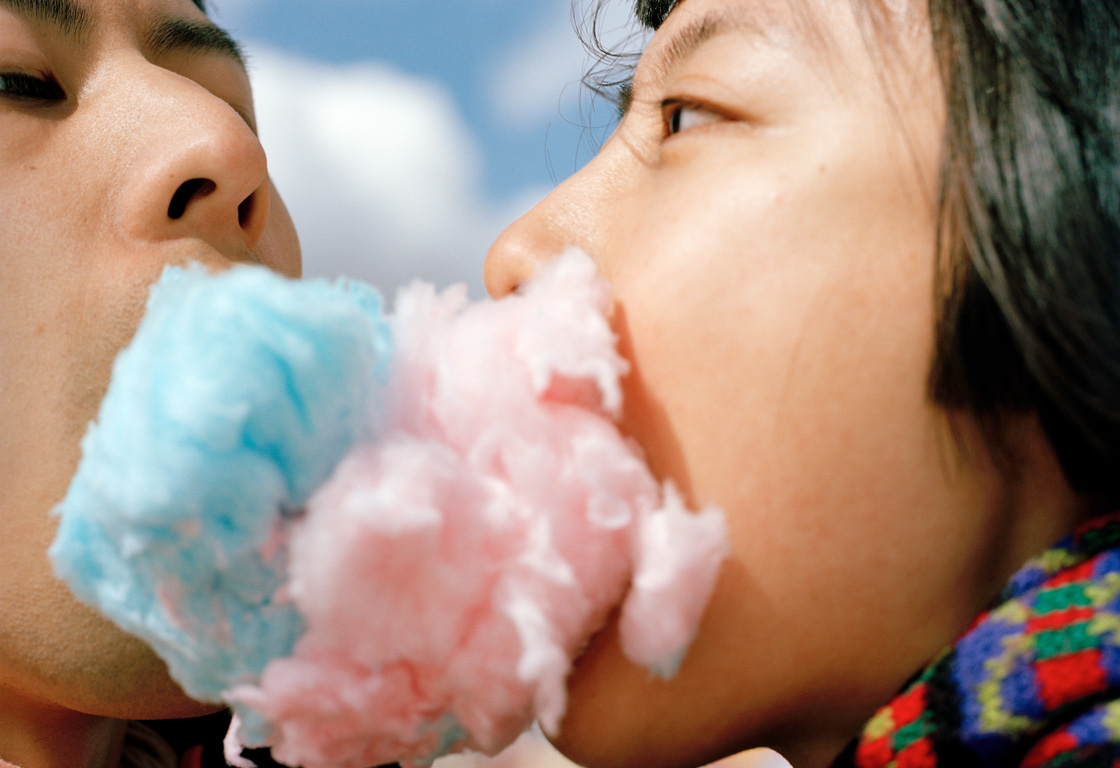 Zwei Personen, die sich nah gegenüberstehen. Die rechte Person hat blaue Zuckerwatte, die linke Person Zuckerwatte im Mund. Die Zuckerwattebäuschchen berühren sich.