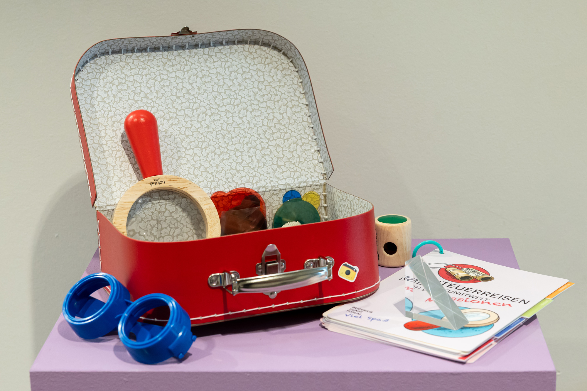A red suitcase stands open on a purple table. To the left of the suitcase are blue glasses. Inside the suitcase is a magnifying glass with a red handle and colourful lenses. To the right of the suitcase is a prism on top of a notebook titled ‘“Abenteuerreisen”' (Adventure Journeys) and a wooden periscope.