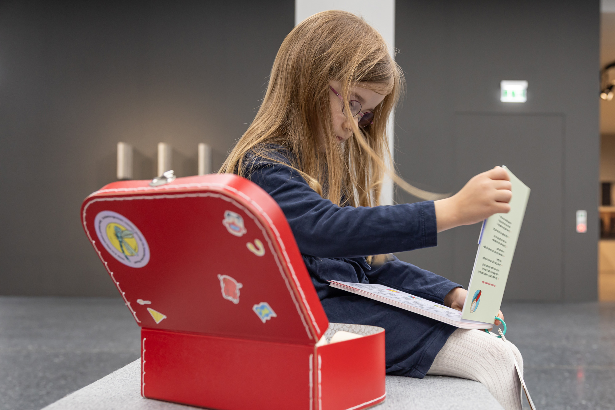 A child sits on a bench leafing through a book. Next to him is an open red suitcase decorated with stickers. The background is dark and shows a modern, minimalist environment.