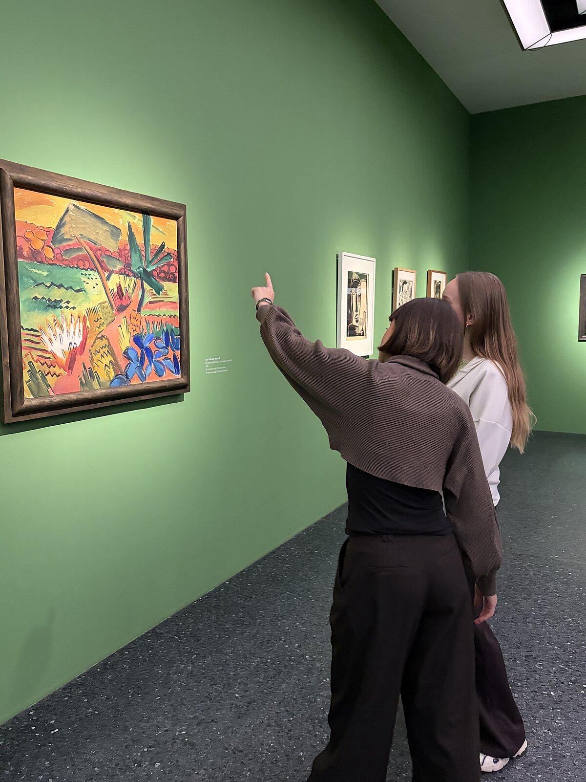 Auf dem Bild sind 2 Personen im Ausstellungraum zu sehen. Sie unterhalten sich und schauen sich Gemälde an.