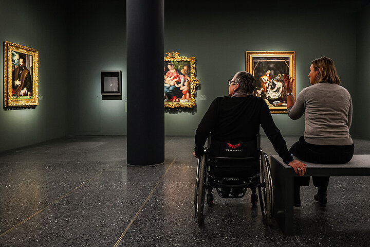 Auf dem Bild sind 2 Personen im Ausstellungraum zu sehen. Sie unterhalten sich und schauen sich Gemälde an.