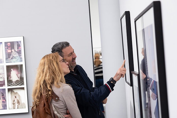 Auf dem Bild sind 2 Personen im Ausstellungraum zu sehen. Sie unterhalten sich und schauen sich Fotografien an.