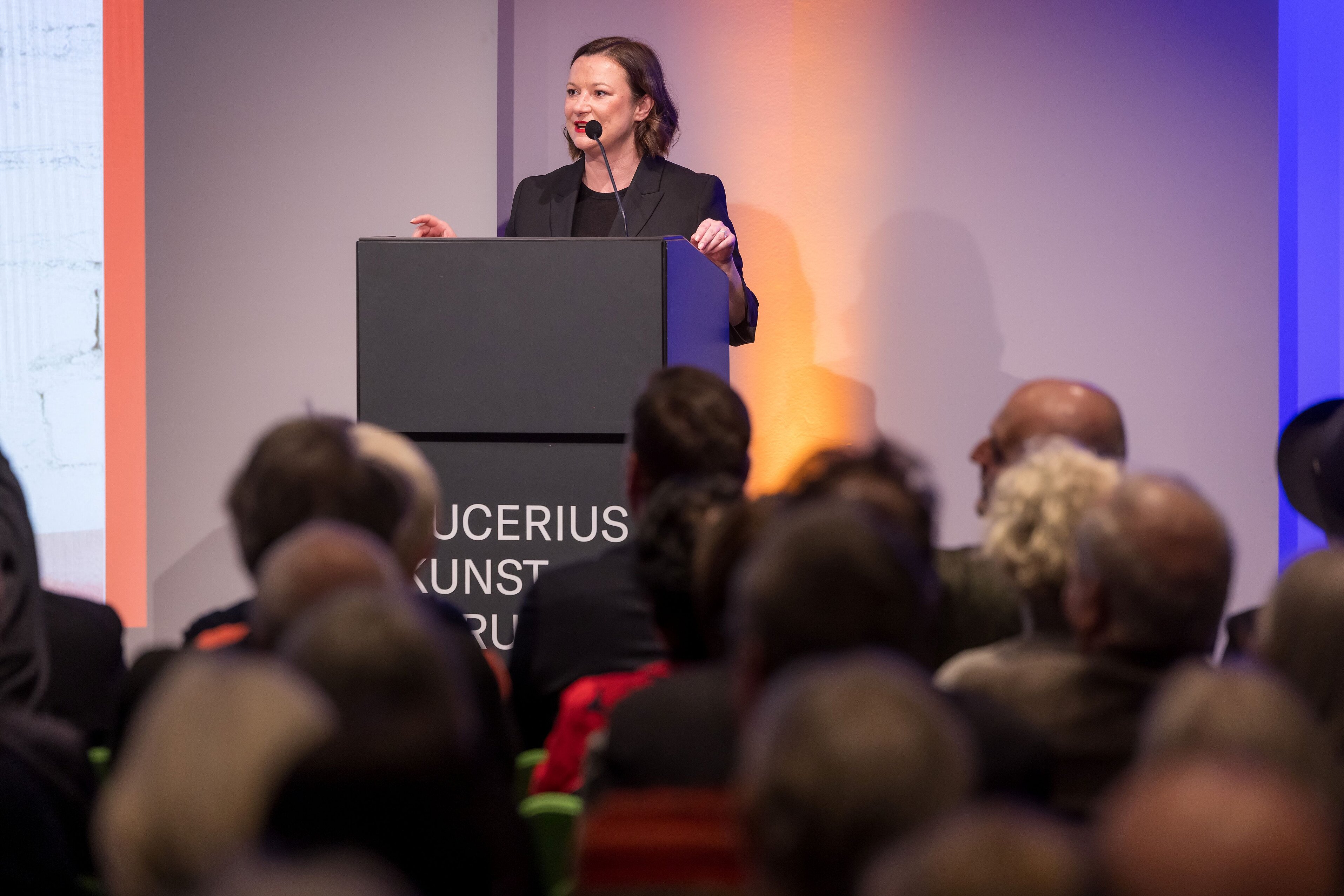 Kathrin Baumstark steht am Rednerpult im Auditorium, vor ihr sitzt Publikum
